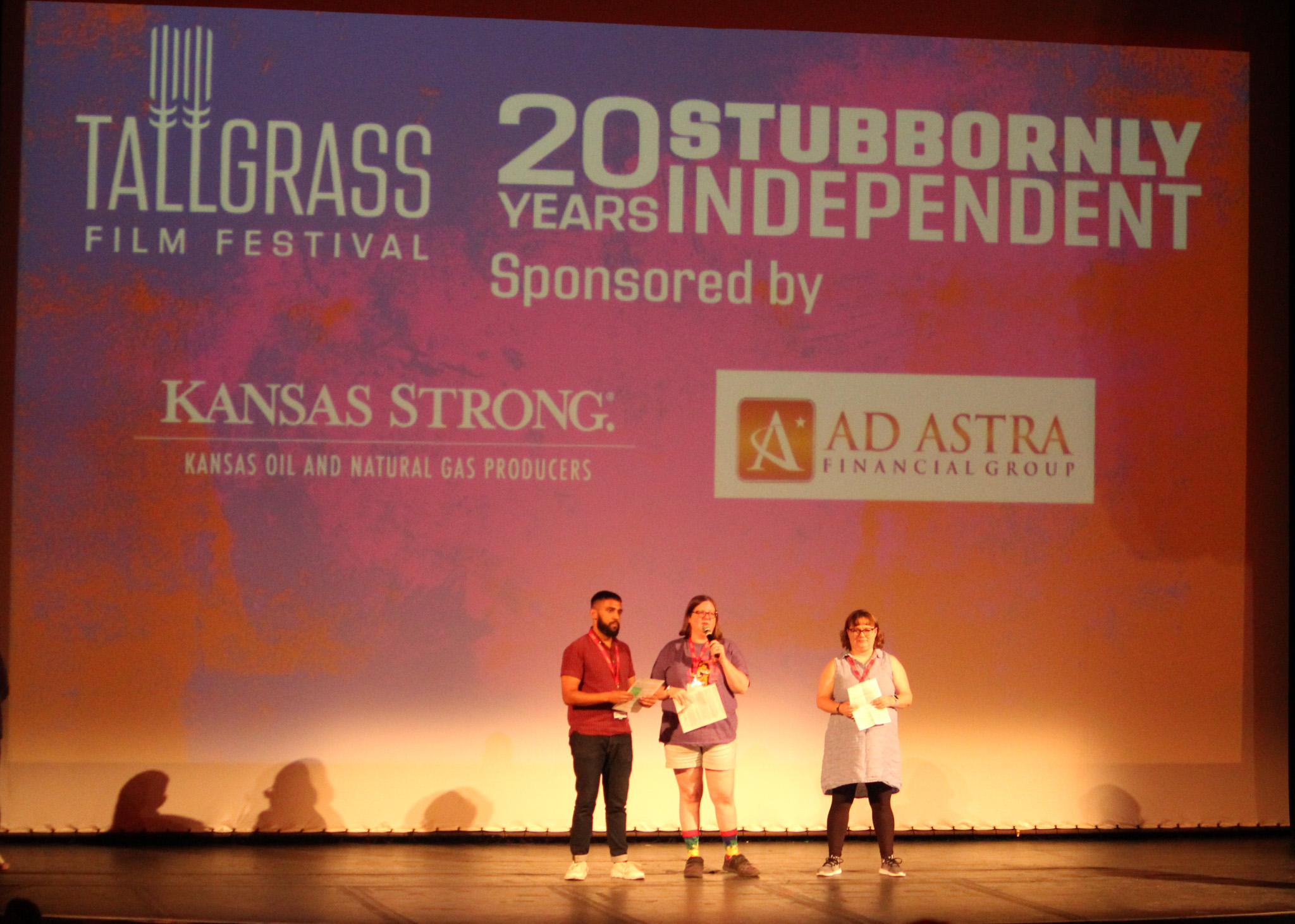 Three people stand on a stage in front of a large screen that has a sponsor logo
