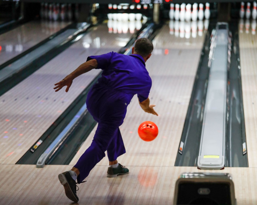 A bowler dressed as Jesus from The Big Lebowski throws an orange bowling ball down a bowling lane