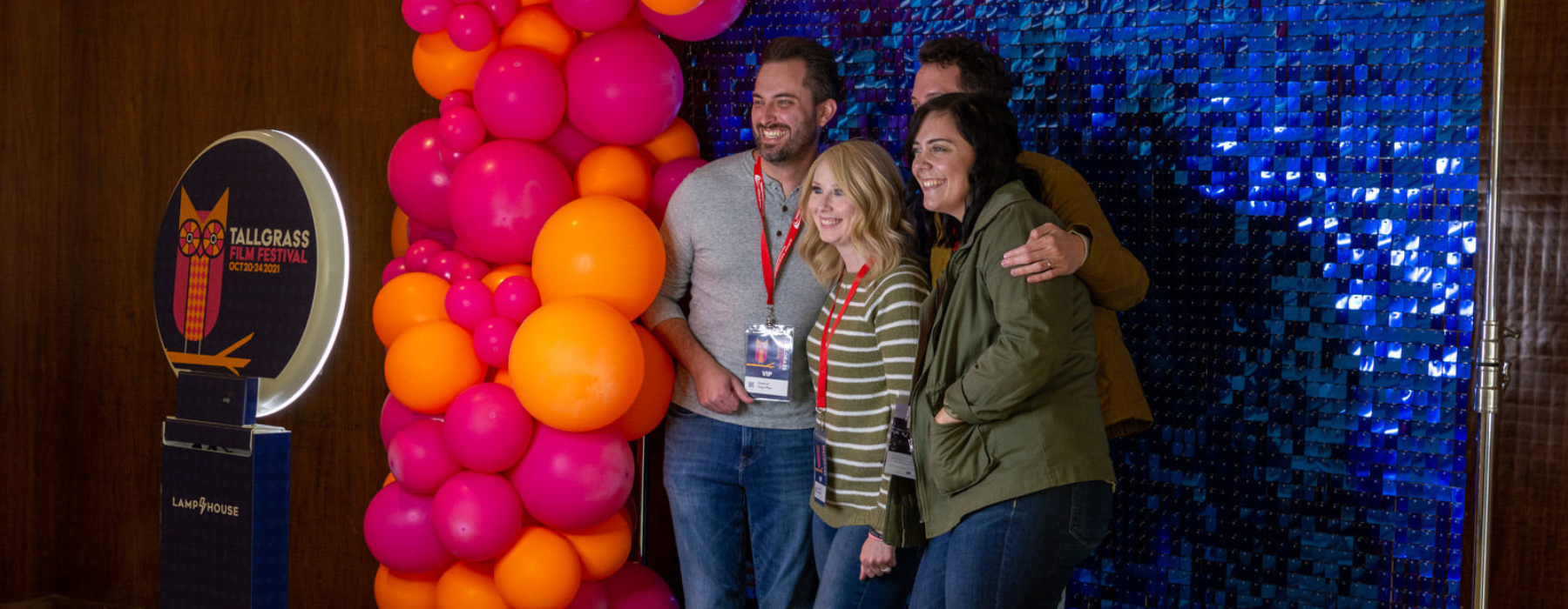 Four people take a picture at a photo booth, there is a orange and pink balloon arch beside them and a shiny blue background behind them