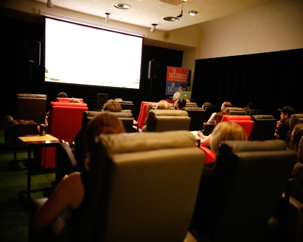 An audience sits in gray, black, and red recliners and looks at a screen and a person giving a presentation