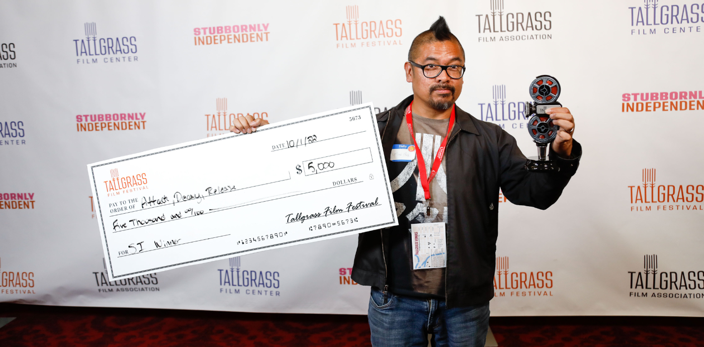 Man with a mohawk holds a large check for Attack Decay Release for 5,000 for the Stubbornly Independent award and a tap handle award in front of a Tallgrass background.