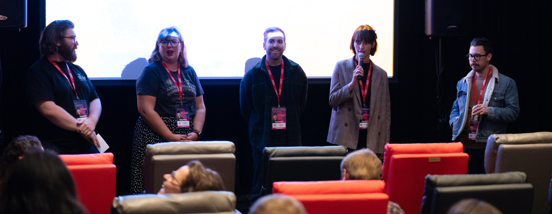 Five people stand in front of a screen facing an audience in red, gray and black chairs