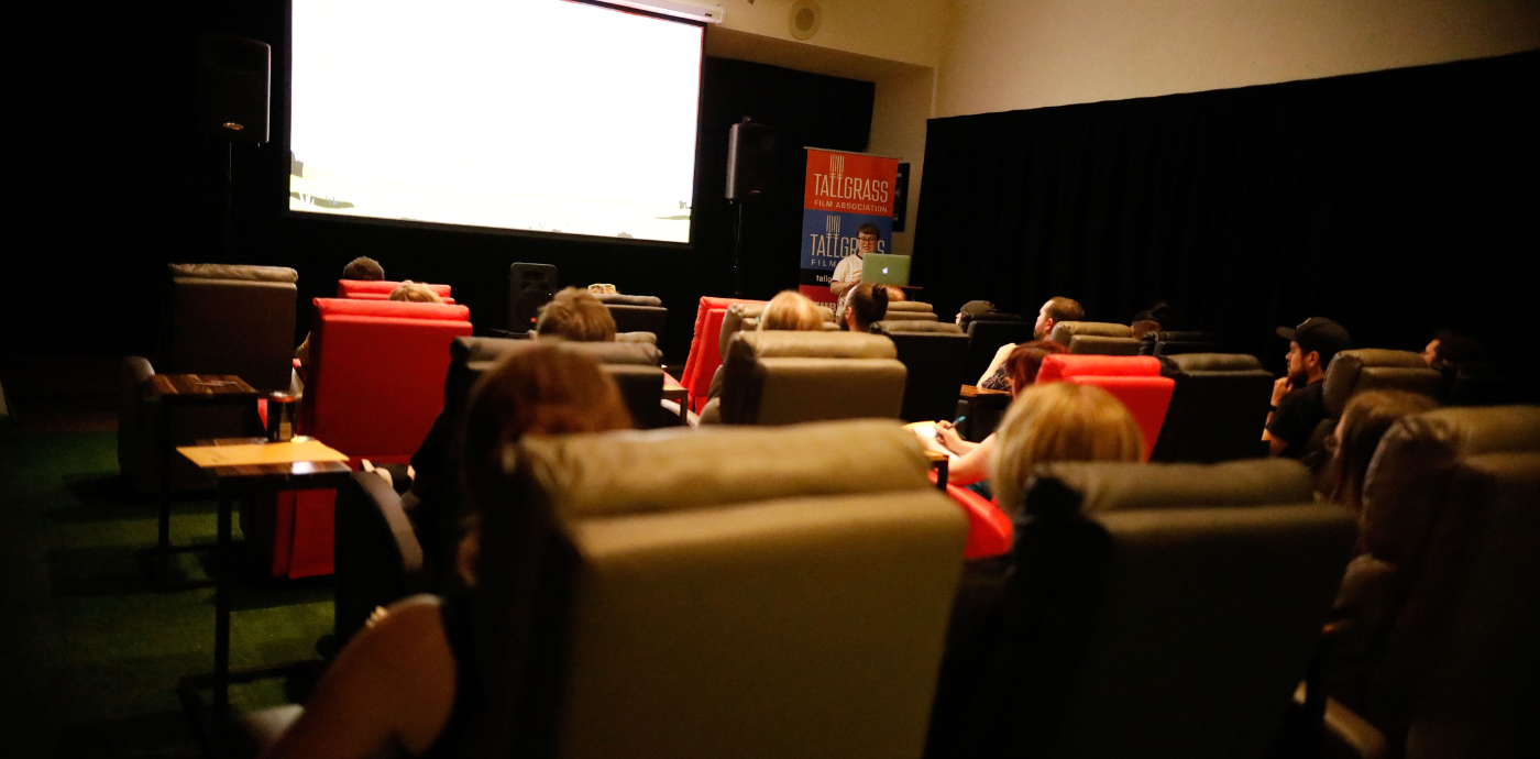An audience sits in recliners watching a screen and someone giving a presentation.