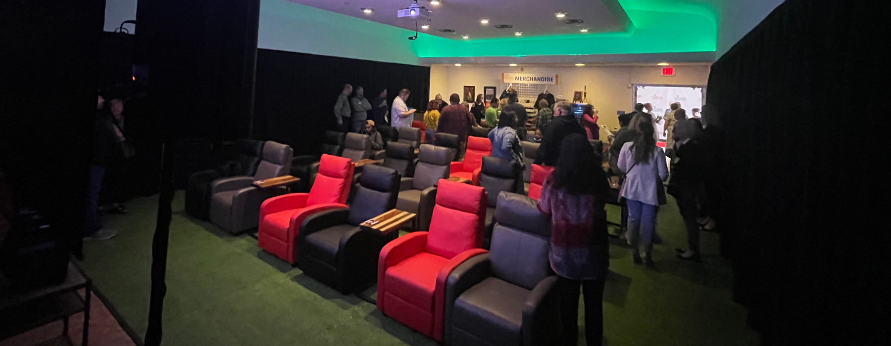 Red, gray and black chairs on green carpet facing a screen, people mingling in the back, green lights light up the ceiling.
