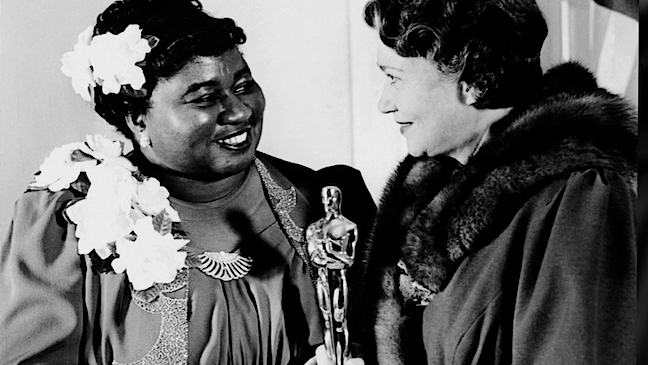 Two women smiling at each other, one holding an Oscar statue, in a vintage black and white photo.